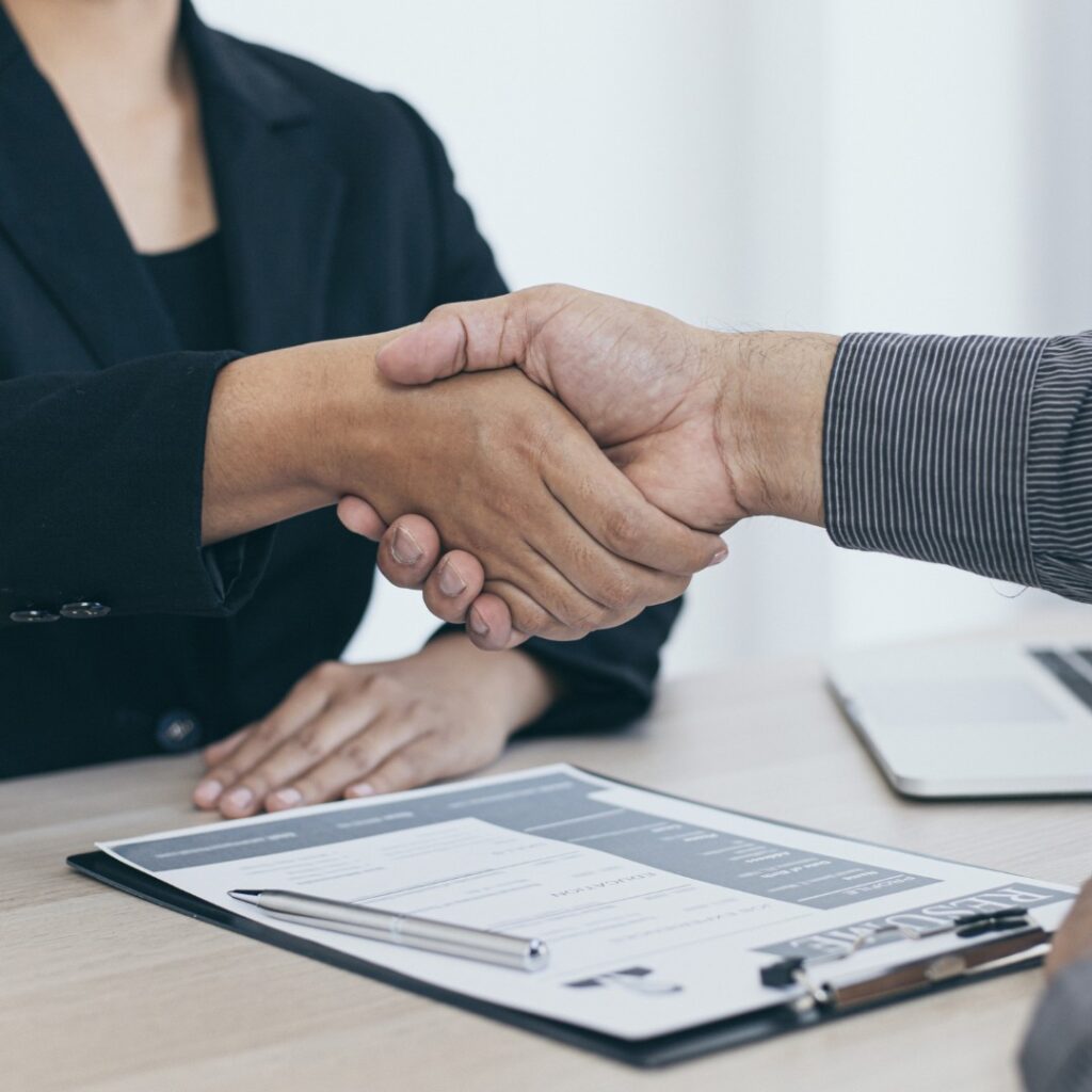 Aperto de mãos entre dois profissionais após uma entrevista de emprego, com currículo e caneta sobre a mesa.