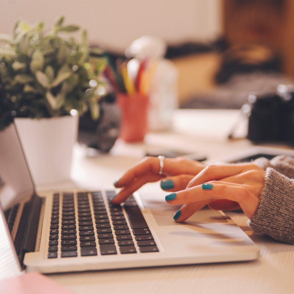 Pessoa digitando em um laptop com unhas pintadas, planta ao lado, em uma mesa de trabalho organizada. Trabalho remoto.