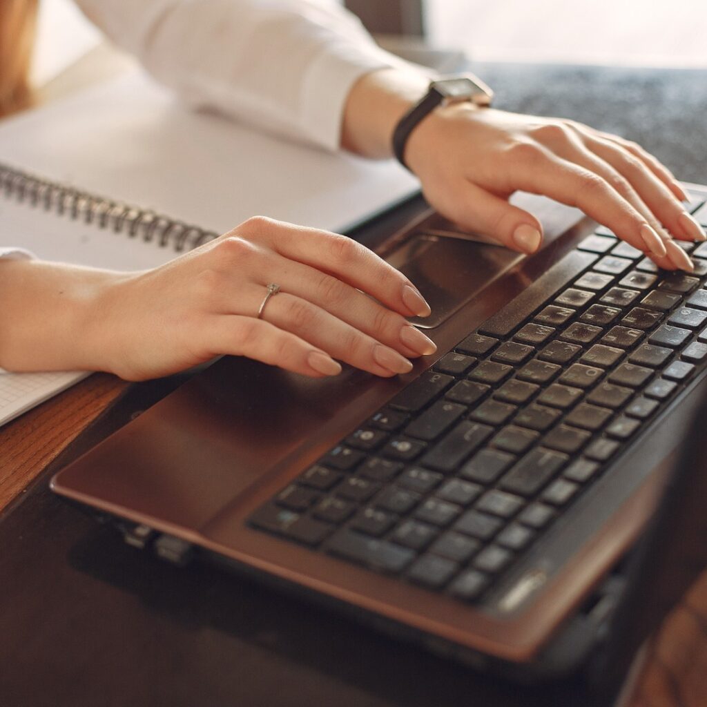 Mãos femininas digitando em um laptop ao lado de um caderno, representando trabalho remoto ou estudo online.