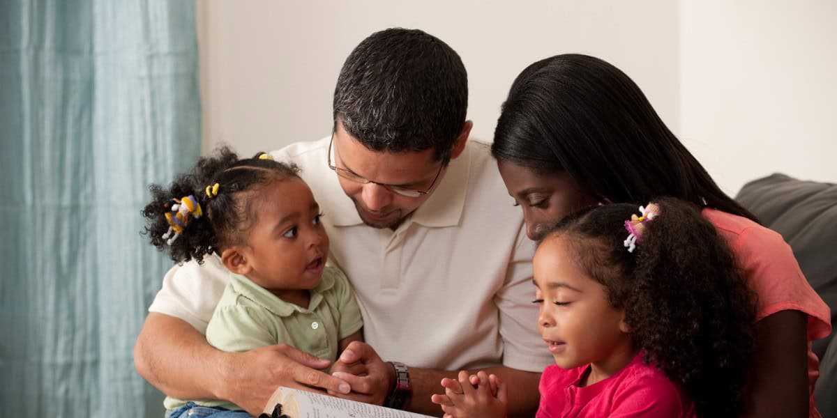 Família reunida lendo livro e orando, pais e duas crianças pequenas em momento de união e espiritualidade.