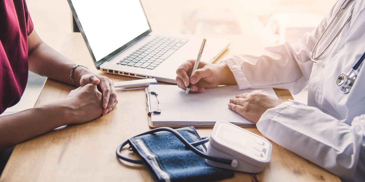 Médico e paciente durante uma consulta médica com computador, prancheta e esfigmomanômetro sobre a mesa.